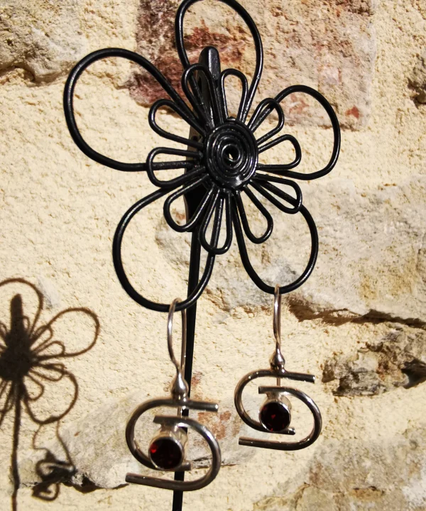 Une boucle d'oreille pendante en argent sterling au design contemporain et architectural, ornée d'un grenat rond et facetté, délicatement portée à l'oreille d'une femme.